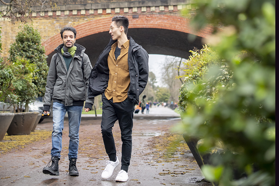 Two male students, wearing warm clothes, walking under a bridge in Ravenscourt Park, surrounded by trees and bushes.